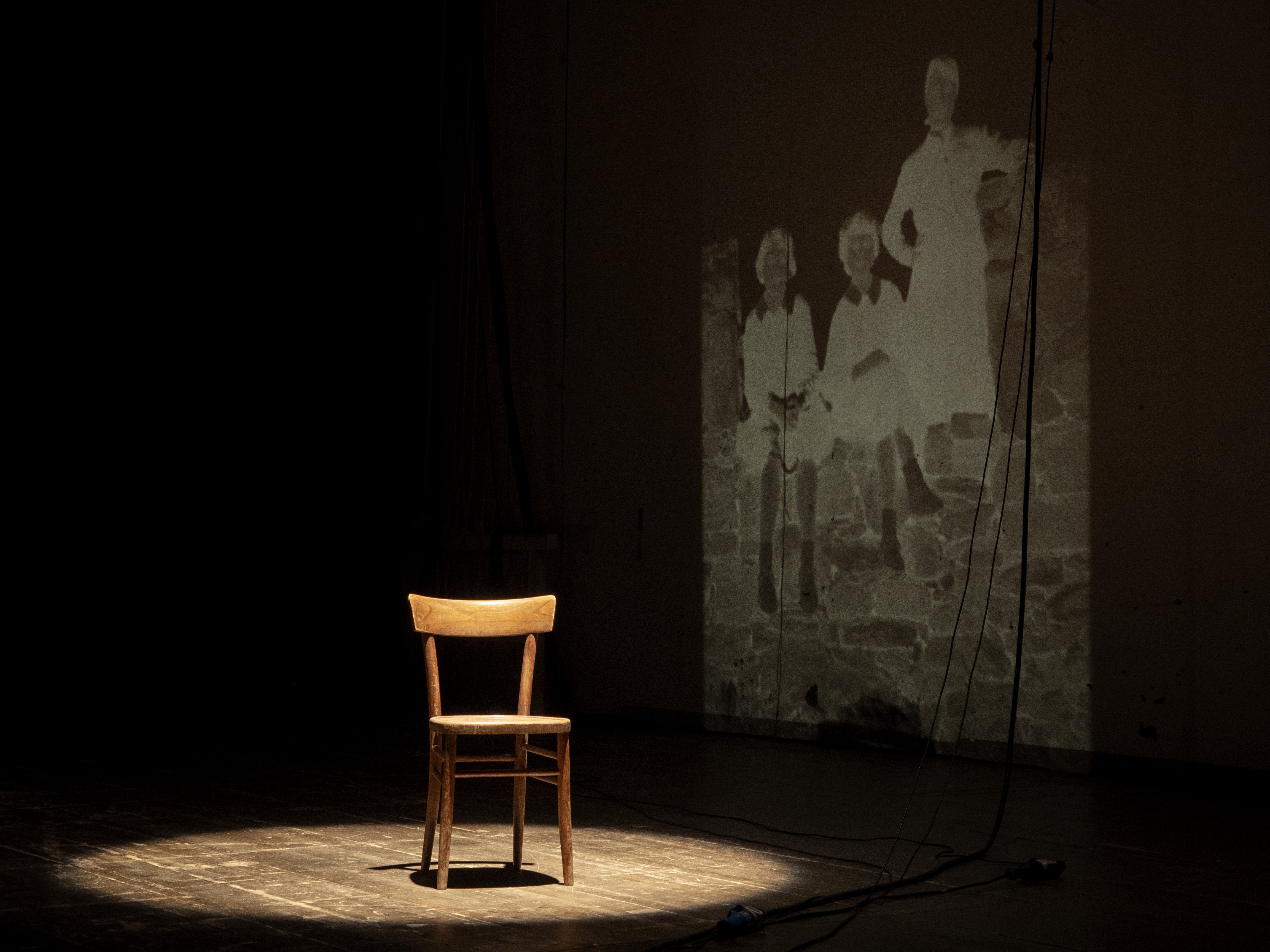 Man sieht einen Holzstuhl auf einer Bühne, der Stuhl ist mit einem Scheinwerfer beleuchtet. Im Hintergrund sieht man die Projektion eines alten Fotos.