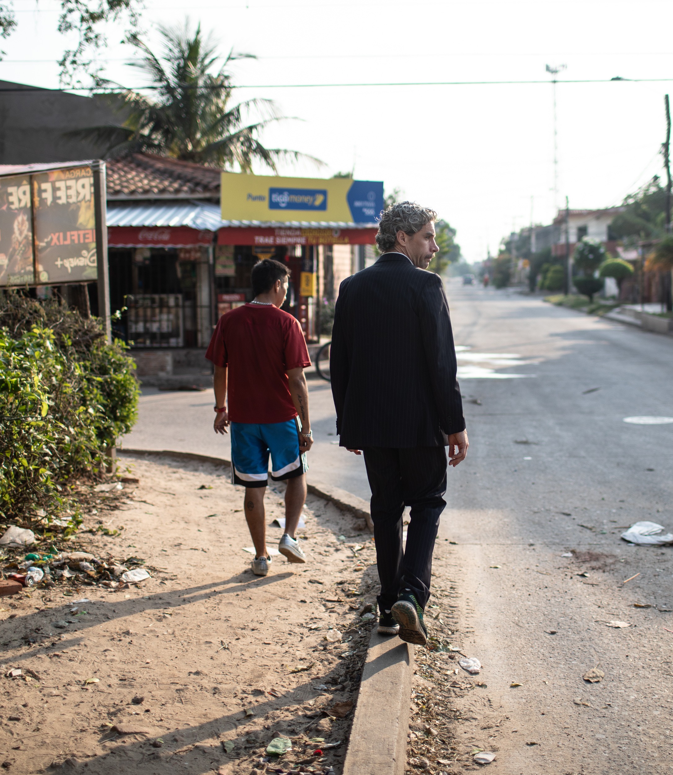 Das Bild wurde draussen aufgenommen. Es zeigt eine staubige Strasse mit ein paar Gebäuden und Pflanzen. Auf der Strasse gehen Jorge Arias Cortez und Nicola Franzen, Nicola im Anzug und Jorge in sportlicher Kleidung.