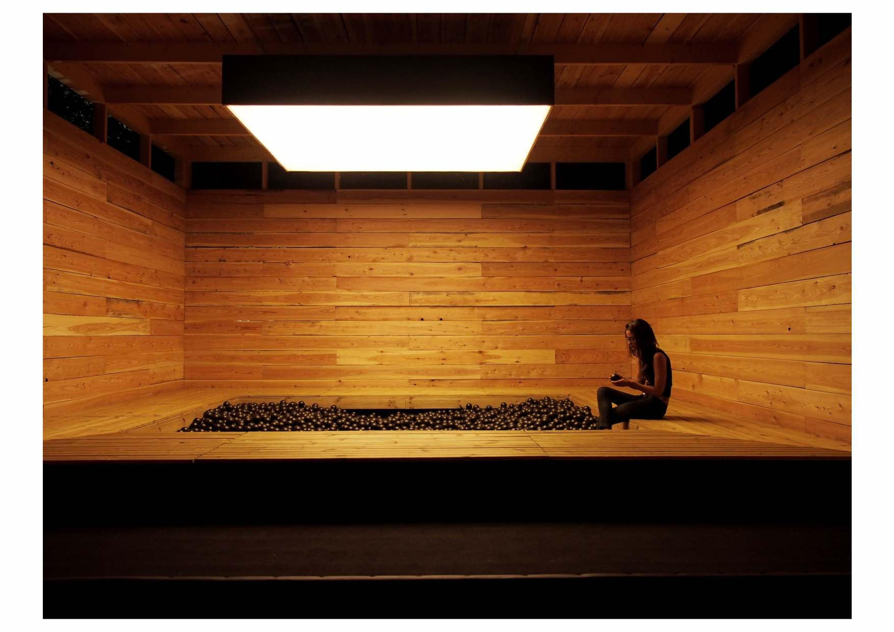 Man sieht in einen Raum aus Holz. In seiner Mitte befindet sich eine Art Swimmingpool, der mit Tausenden von schwarzen Plastikbällen gefüllt ist. Am rechten Rand des Pools sitzt eine Person.